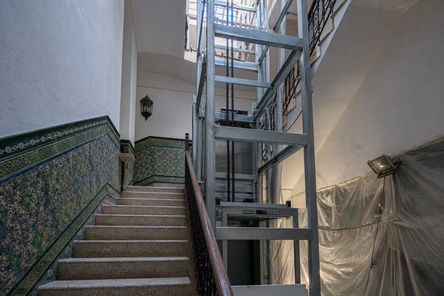 Hueco de escalera de una comunidad con zócalo de azulejo y una estructura metálica de ascensor a medio montar