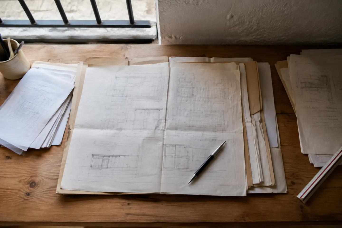 Carpeta de expediente abierta con planos acotados y copias de escritura sobre una mesa junto a una ventana con reja