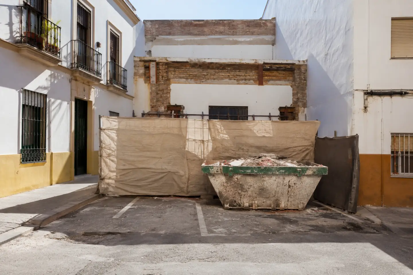 Contenedor de escombro y valla de obra ocupando una plaza de aparcamiento en una calle estrecha de Sevilla