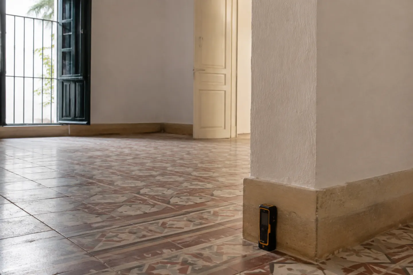 Distanciómetro láser apoyado en el rodapié de una estancia vacía con la solería hidráulica a la vista