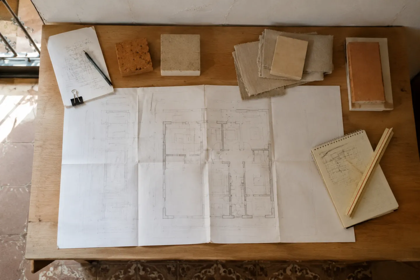 Mesa de trabajo con plano de una vivienda, muestras neutras de materiales y notas de distribución