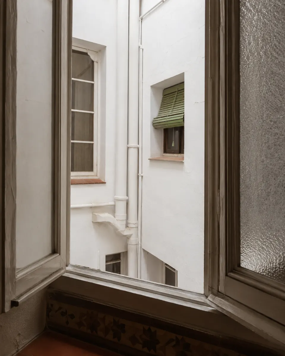 Patio de luces de una casa de pisos sevillana visto desde una ventana interior, con bajantes vistos y la pared encalada