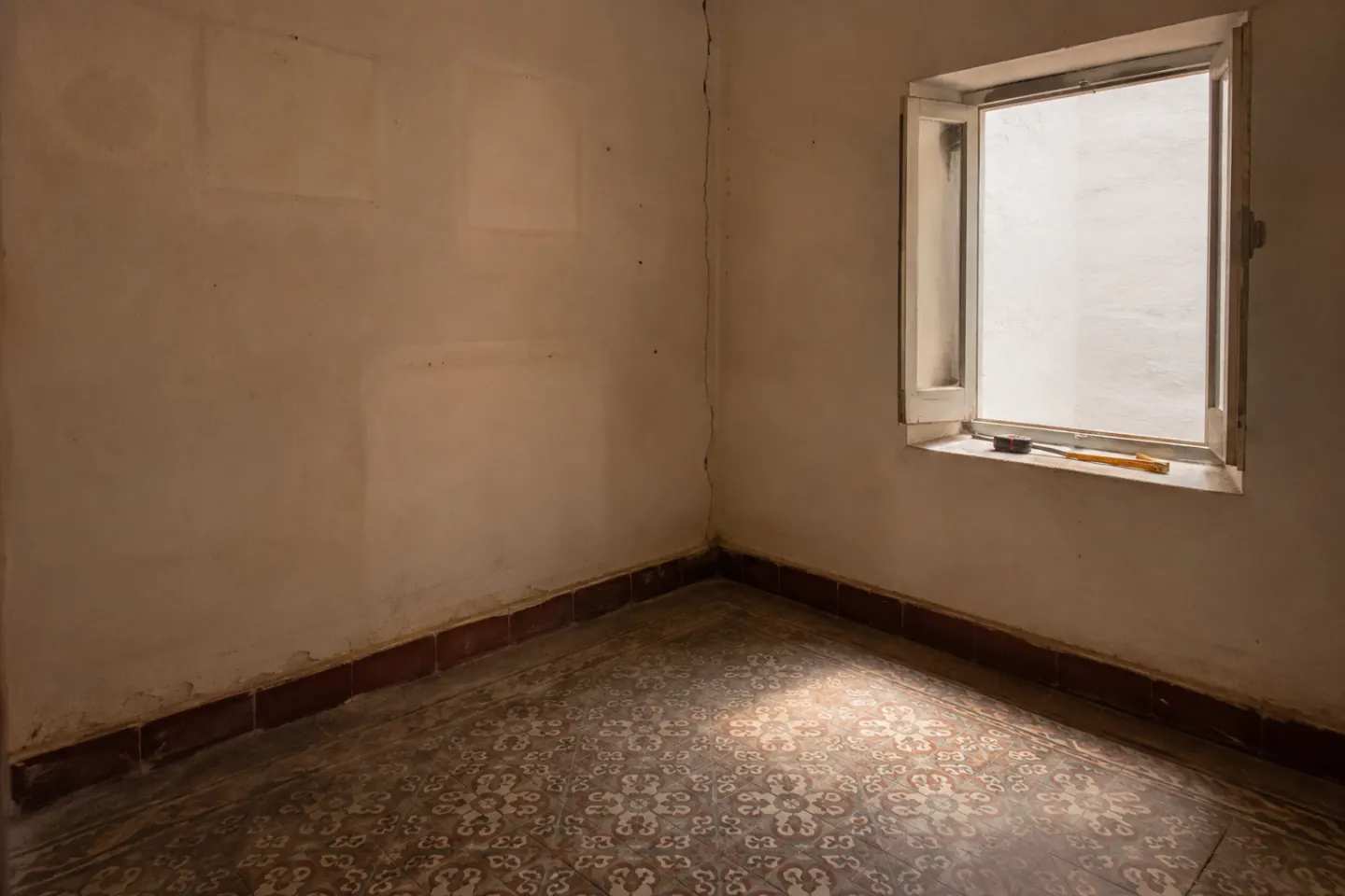 Piso vacío con suelo hidráulico y una persiana entreabierta, con un medidor láser sobre el alféizar